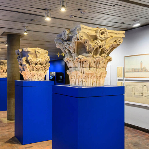 A long view of replical column capitals on pedestals in front of a wall of images.