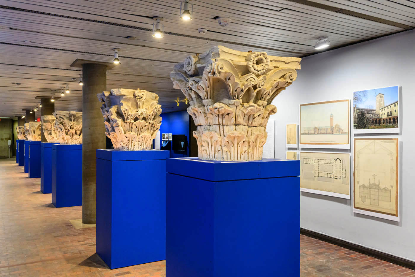 A long view of replical column capitals on pedestals in front of a wall of images.