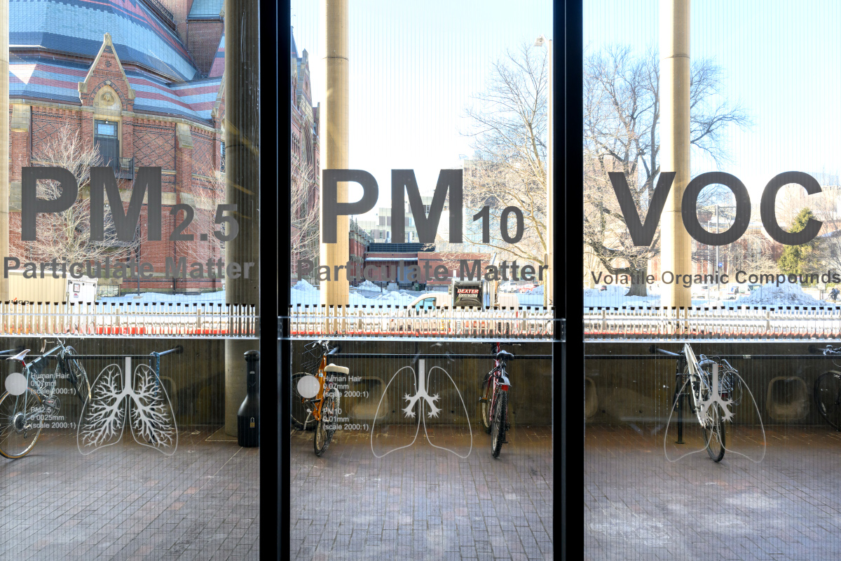 A collection of test tubes displayed in the windows of Frances Loeb Library beneath the words, “Particulate Matter” and “Volatile Organic Compounds.”