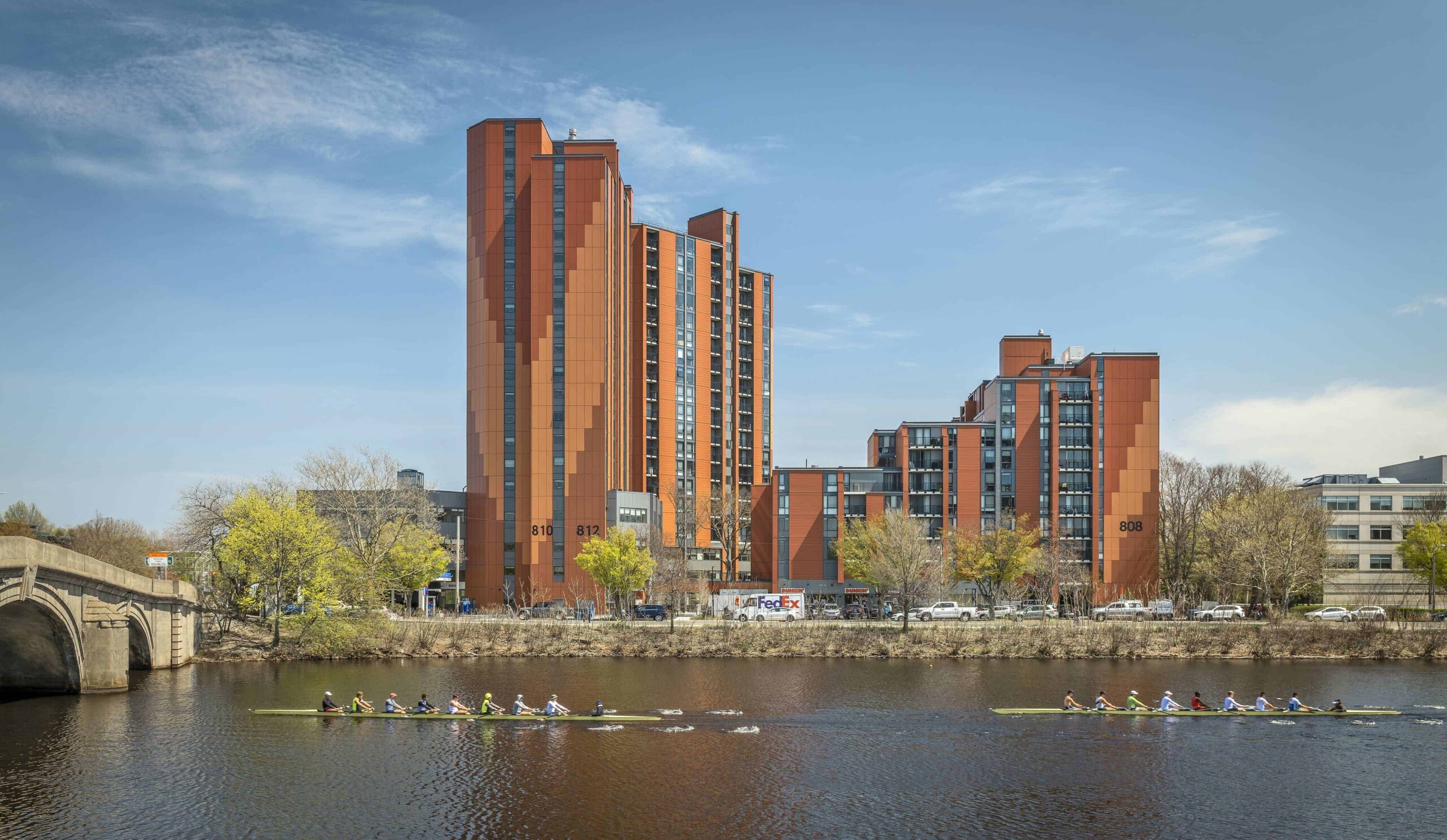 Photograph of tall terra cotta apartment building behind rowers on the Charles river