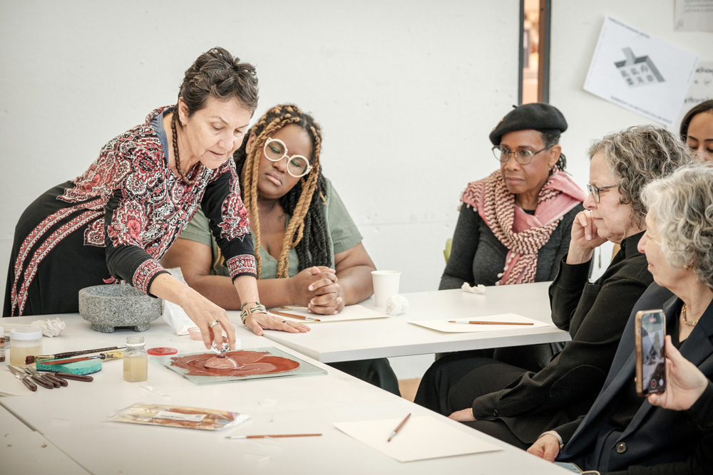 A photograph showing an artist pointing to a design on a sheet of paper while women seated at the table look on.