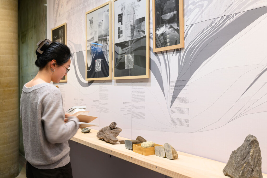 A person looking at framed drawings against a wall sized mural of a geologic section of massachusetts. With a shelf in front with rock on top.