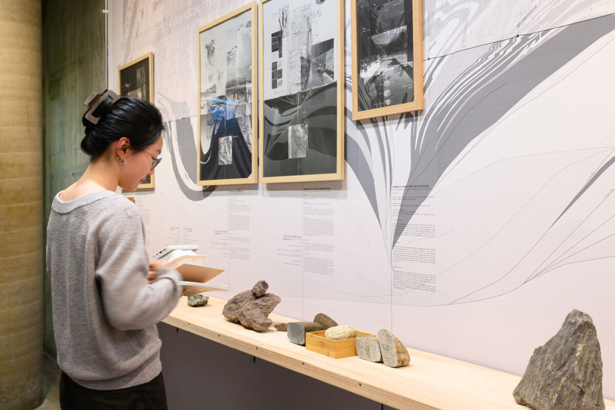 A person looking at framed drawings against a wall sized mural of a geologic section of massachusetts. With a shelf in front with rock on top.