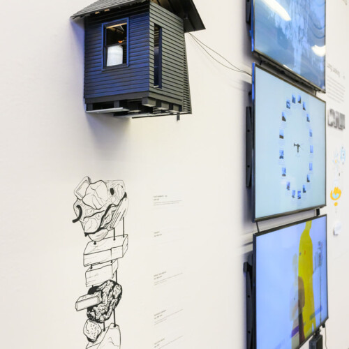 Exhibition wall, showing a detail of a small model of a house directly on top of a hand-drawn geologic section, and three monitors