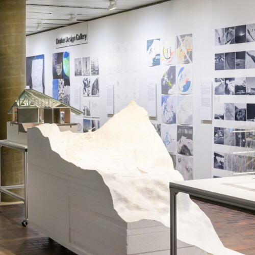 A view of the exhibit in Druker Design Gallery, with a line of tables displaying 3D models and a long wall displaying a variety of 2D images.