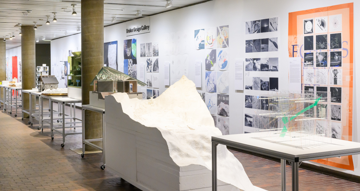A view of the exhibit in Druker Design Gallery, with a line of tables displaying 3D models and a long wall displaying a variety of 2D images.