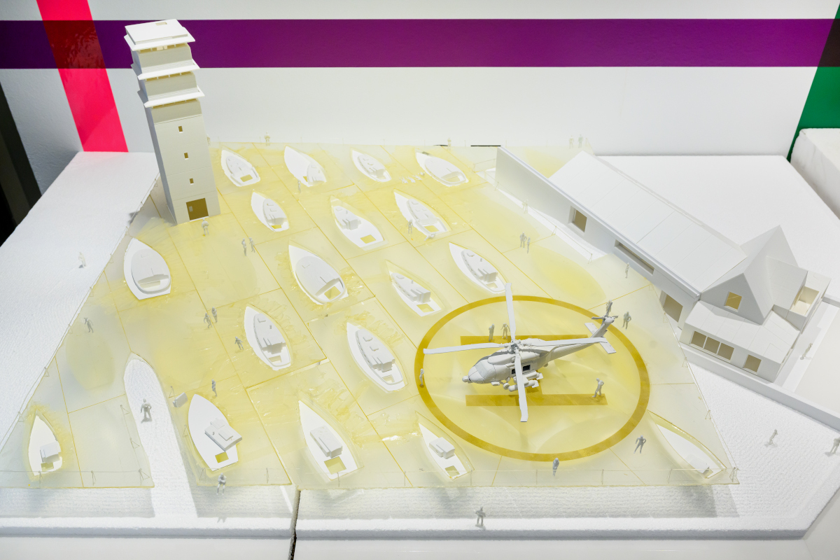 A detail of a 3D model with a helicopter and boats in front of two buildings.