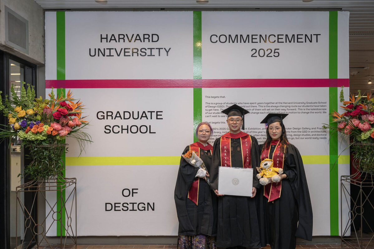 Three recent graduates stand infront of a wall designed with bright colored plaid, flanked by flowers