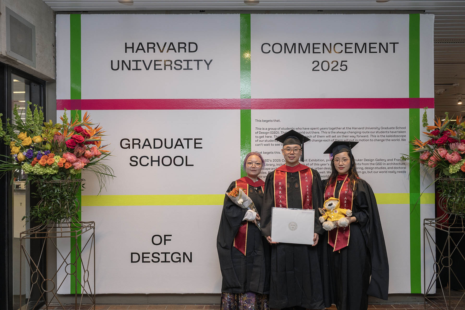 Three recent graduates stand infront of a wall designed with bright colored plaid, flanked by flowers