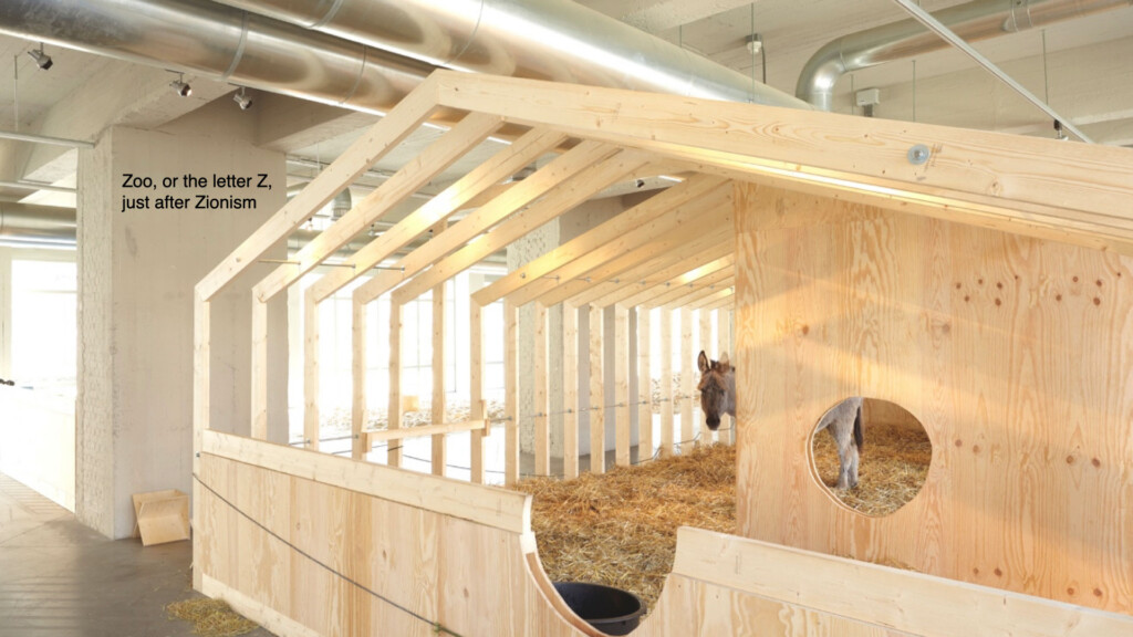 A photograph of a wooden barn structure inside of which is a mule. Text on the wall reads "Zoo, or the letter Z, just after Zionism."