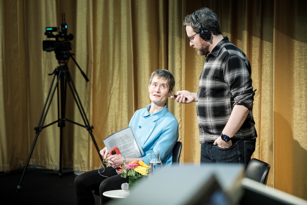 A woman seated on stage holds a book and looks up at a man standing next to her, who is adjusting her microphone. A camera on a tripod stands in the background, and a bouquet of flowers sits on a nearby table.