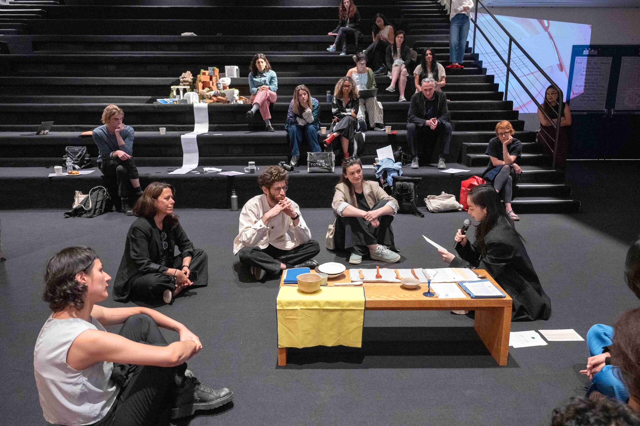 Five students and faculty sit on the floor around a short table in discussion as others watch in the background
