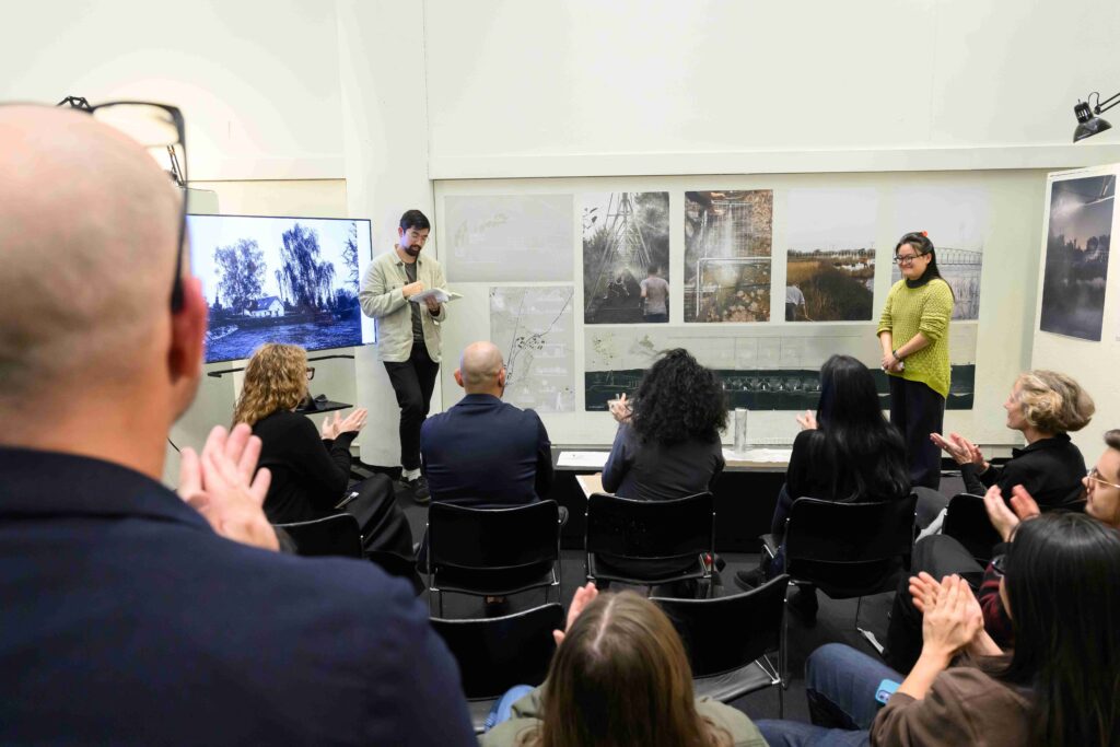 two students stand in front of drawings pinned to a wall and receive applause