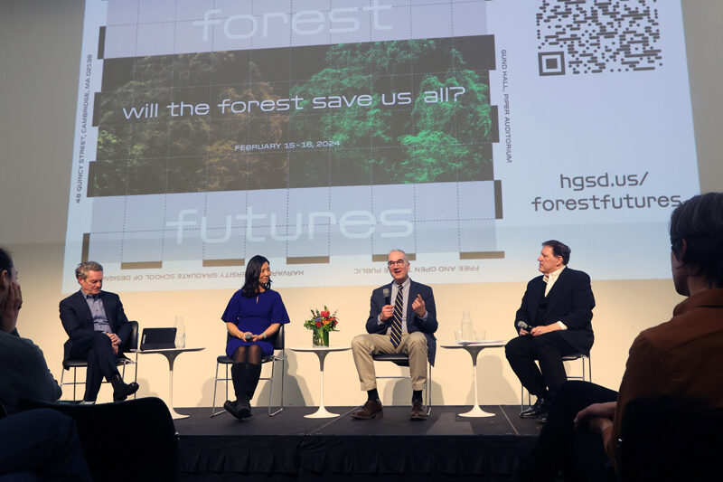 Four people sit on chairs on a low stage as part of a panel discussion. An image projected behind them says Forest Futures: Will the Forest Save us All"