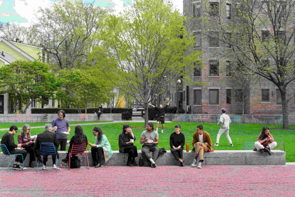 People sit together on a ledge foregrounding a green lawn, where others sit or play volleyball.