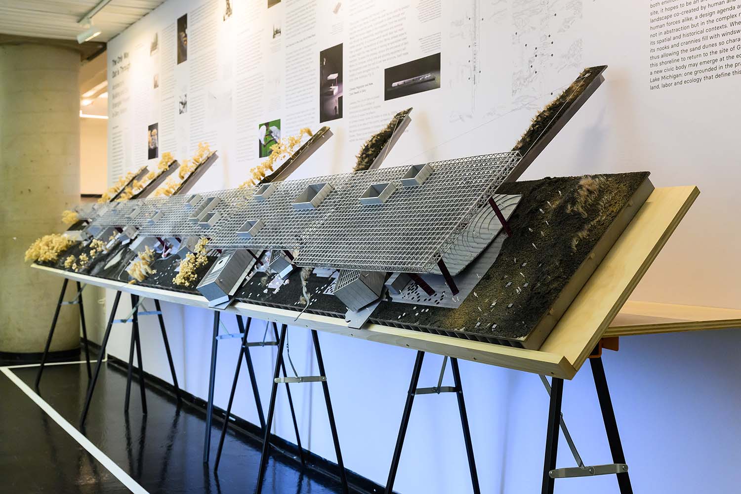 Overview of the exhibition wall of sparse text and images with long tilted model in front of it.
