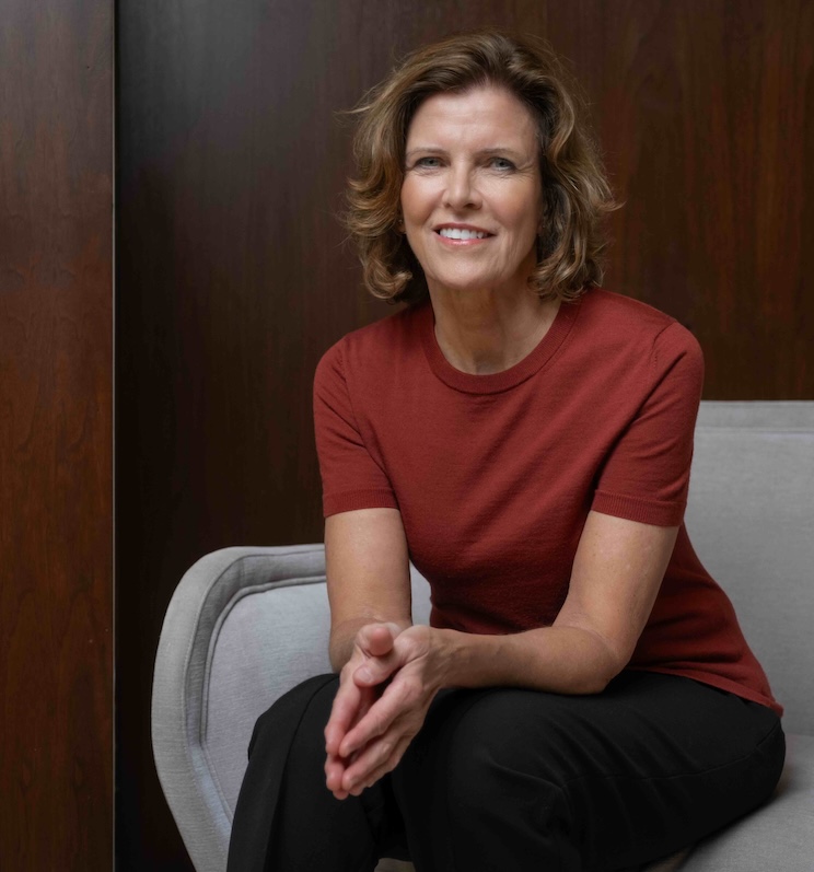 Jeanne Gang, wearing a rust-colored short-sleeve top and black pants, sits on a light gray chair against a dark wooden background, smiling warmly with hands clasped.