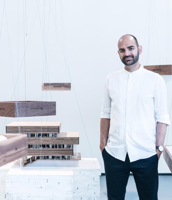 Neeraj Bhatia, a man with a beard and short hair, dressed in a white shirt and black pants, stands indoors next to wooden architectural models suspended by wires. He is smiling slightly and has his hands in his pockets.