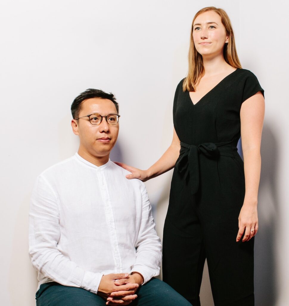 Lap Chi Kwong sits on a stool, wearing a white shirt and glasses. Alison Von Glinow stands next to him, gently resting her hand on his shoulder. She is dressed in a black jumpsuit. Both are looking ahead with neutral expressions, against a plain white background.