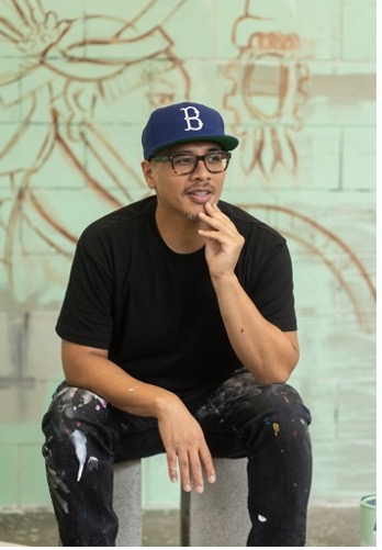 Headshot of Patrick Martinez seated in a chair with his hand placed on his chin against an art mural.