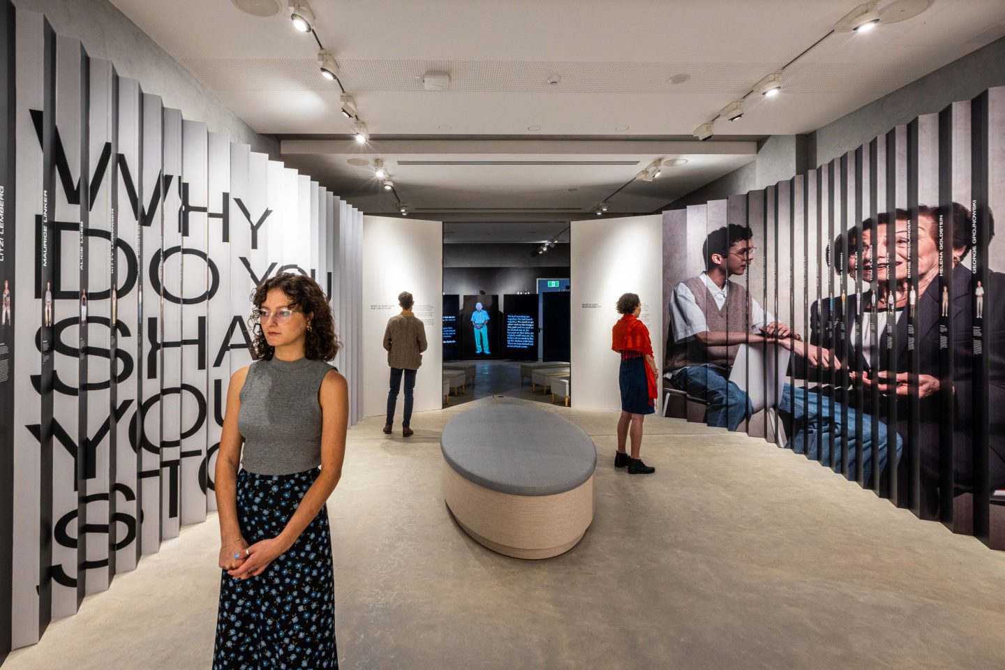 A modern museum exhibit space featuring interactive displays and large vertical panels with text and images. Visitors are walking and observing the exhibit, which includes a bold black text installation reading “WHY DO YOU SHARE YOUR STORY?” and photographic portraits on the walls.