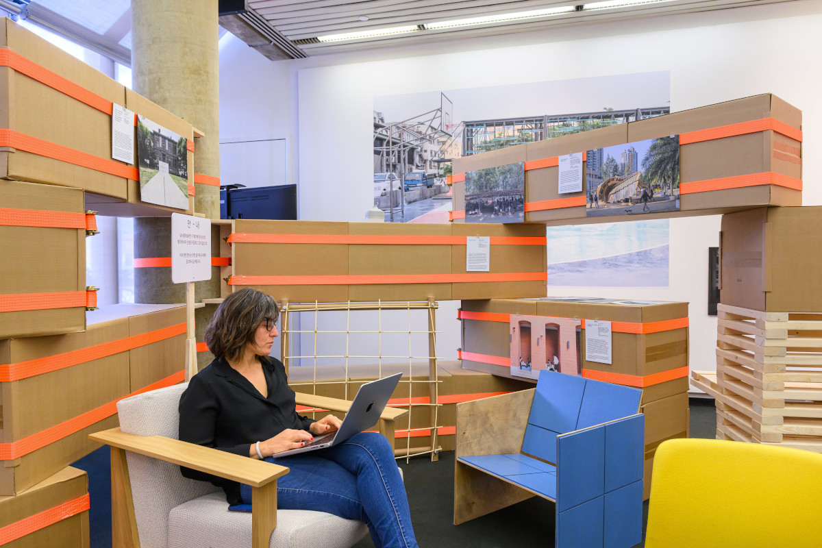 A person seated in an alcove constructed with cardboard boxes and ratchet straps.