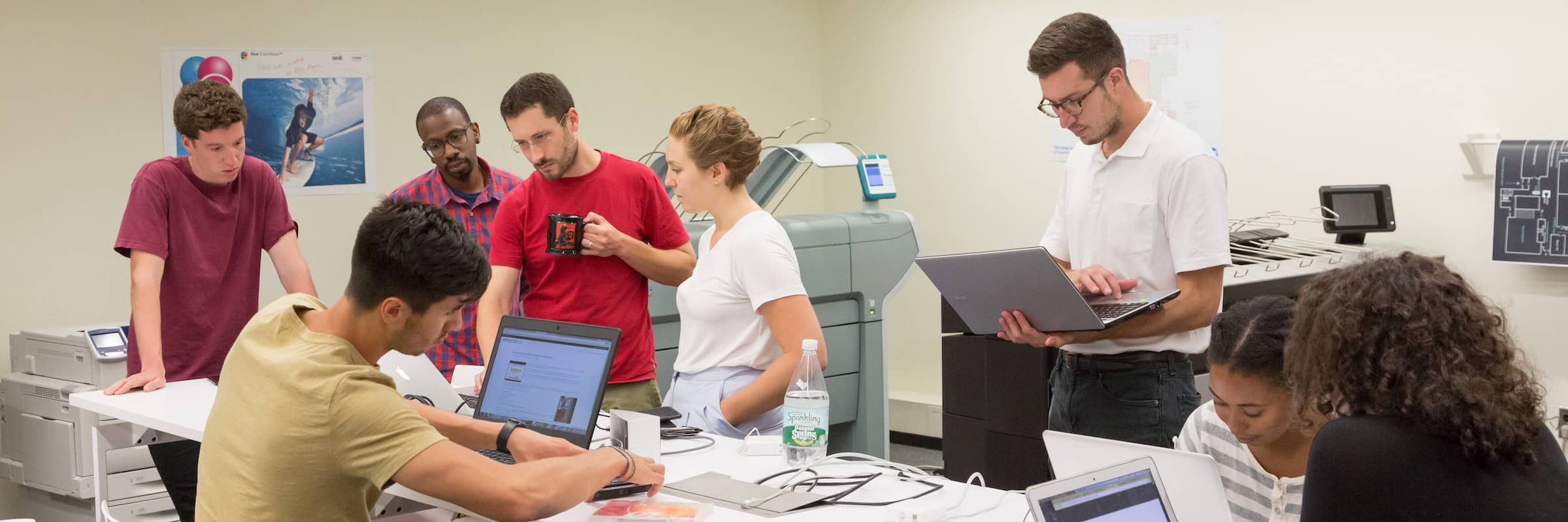 Several people gather around a table while working on laptops.