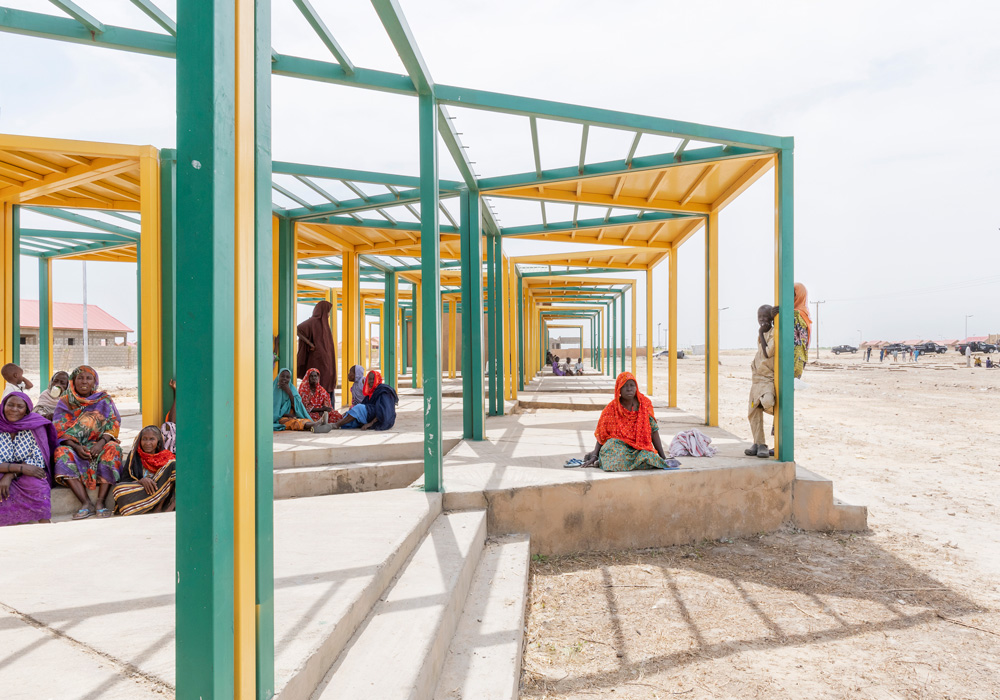 Women in desert-like environment sitting under  green and yellow framework.