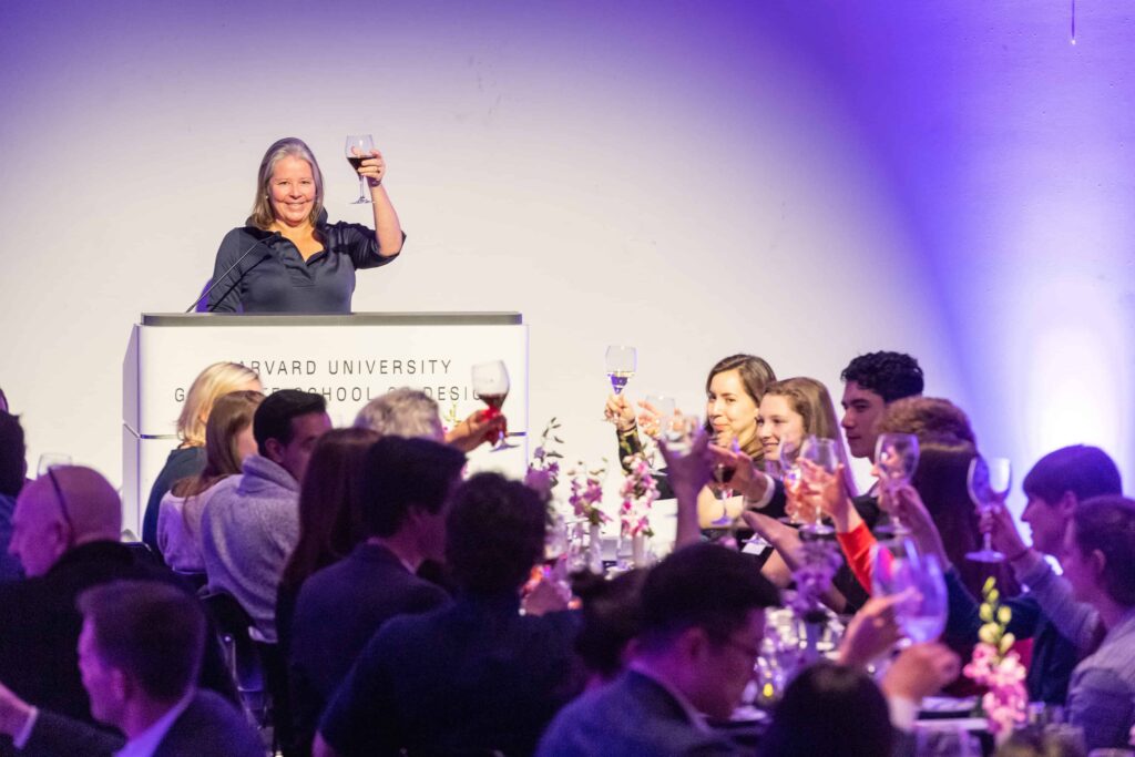 A woman holds up a wine glass at a podium for a toast with a table of people in front of her
