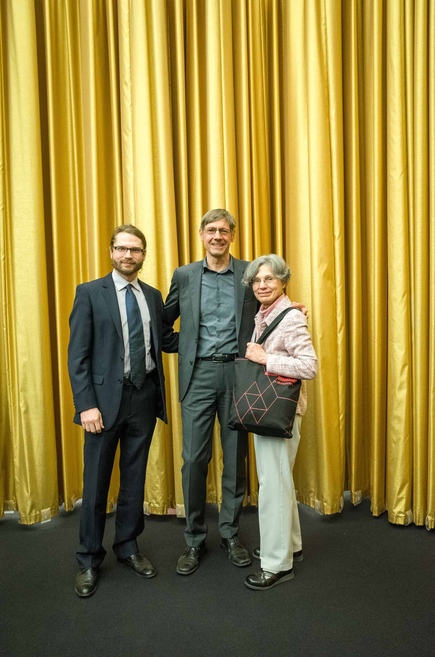 Ben Schodek, Martin Bechthold, and Kay Schodek stand smiling in front of a gold curtain