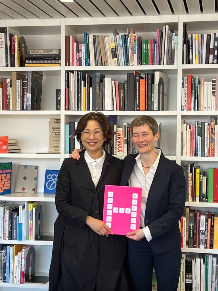 Donor Hailim Suh stands next to Dean Sarah Whiting holding a pink folder with a white H in front of a bookshelf