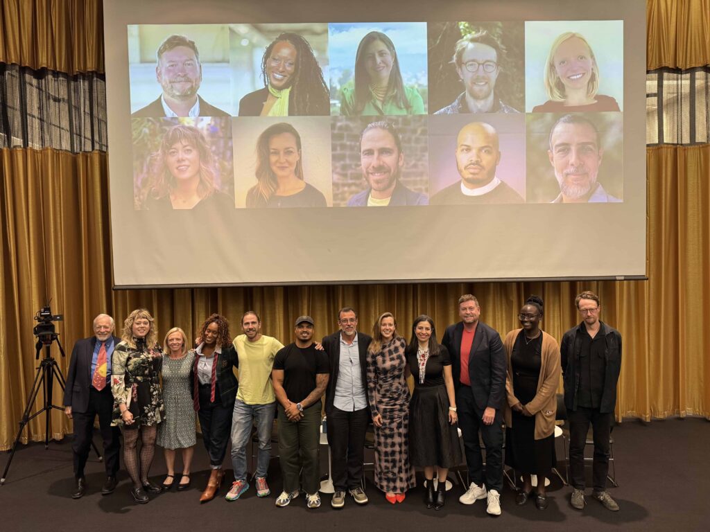 A group of Loeb Fellows Class of 2026 stand in front of a projection grid of their headshots