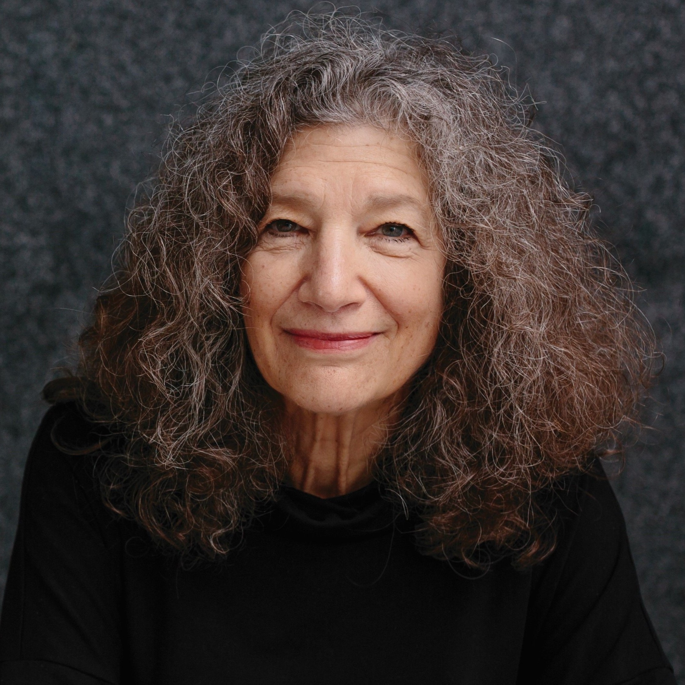 A woman with curly hair smiles in front of a grey background.