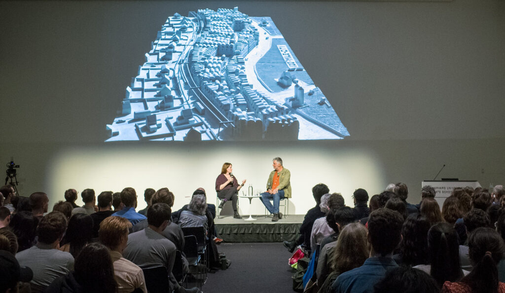 Two people talk on a platform in front of an audience, with a projects housing model in the background.