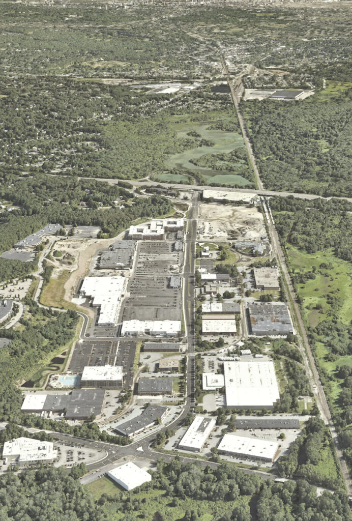 Aerial view of a group of single story industrial buildings and parking lots surrounded by a green wooded landscape.