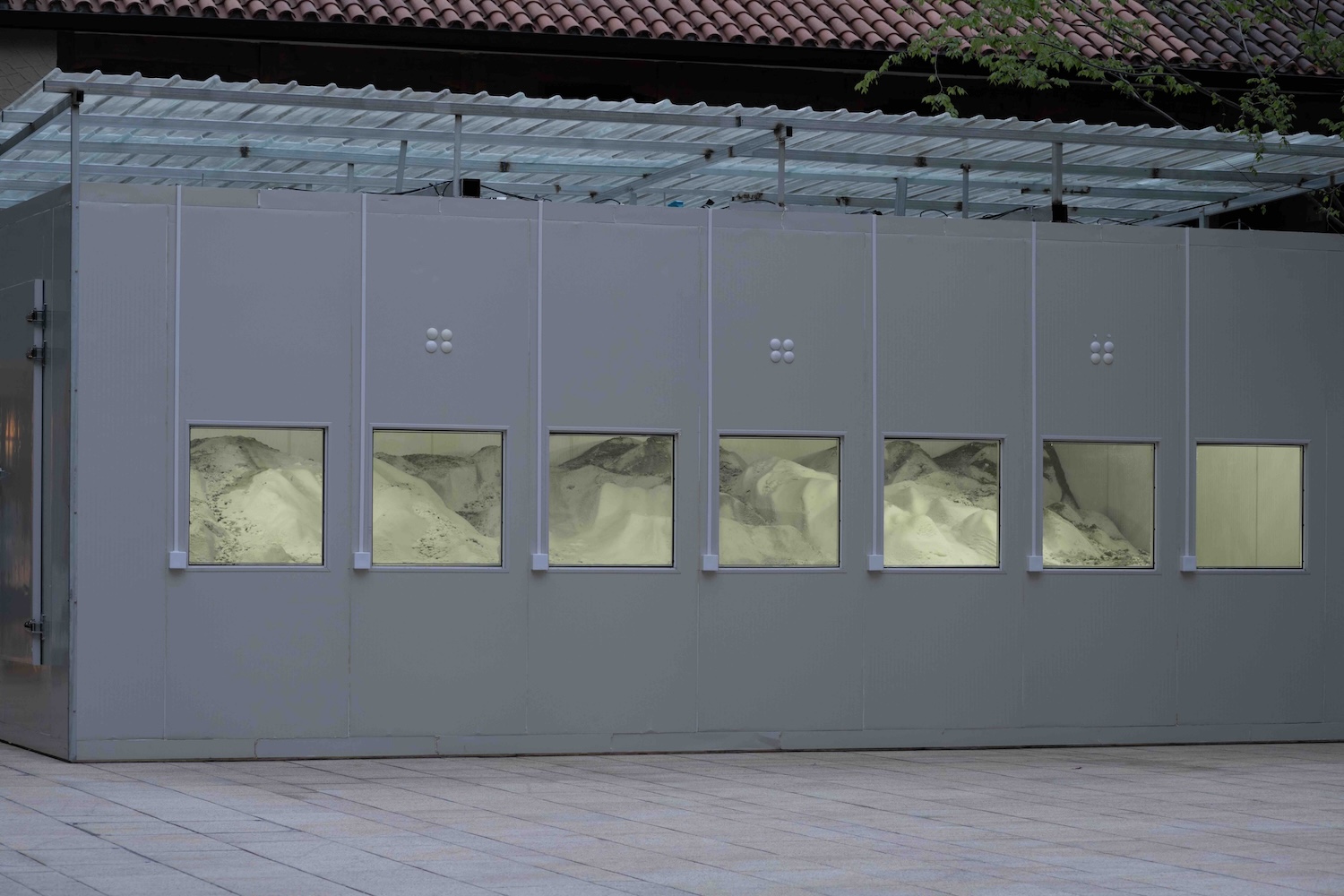 Grey industrial-looking structure with six lit windows revealing mounded, pale material or sand inside, set in an outdoor paved courtyard.