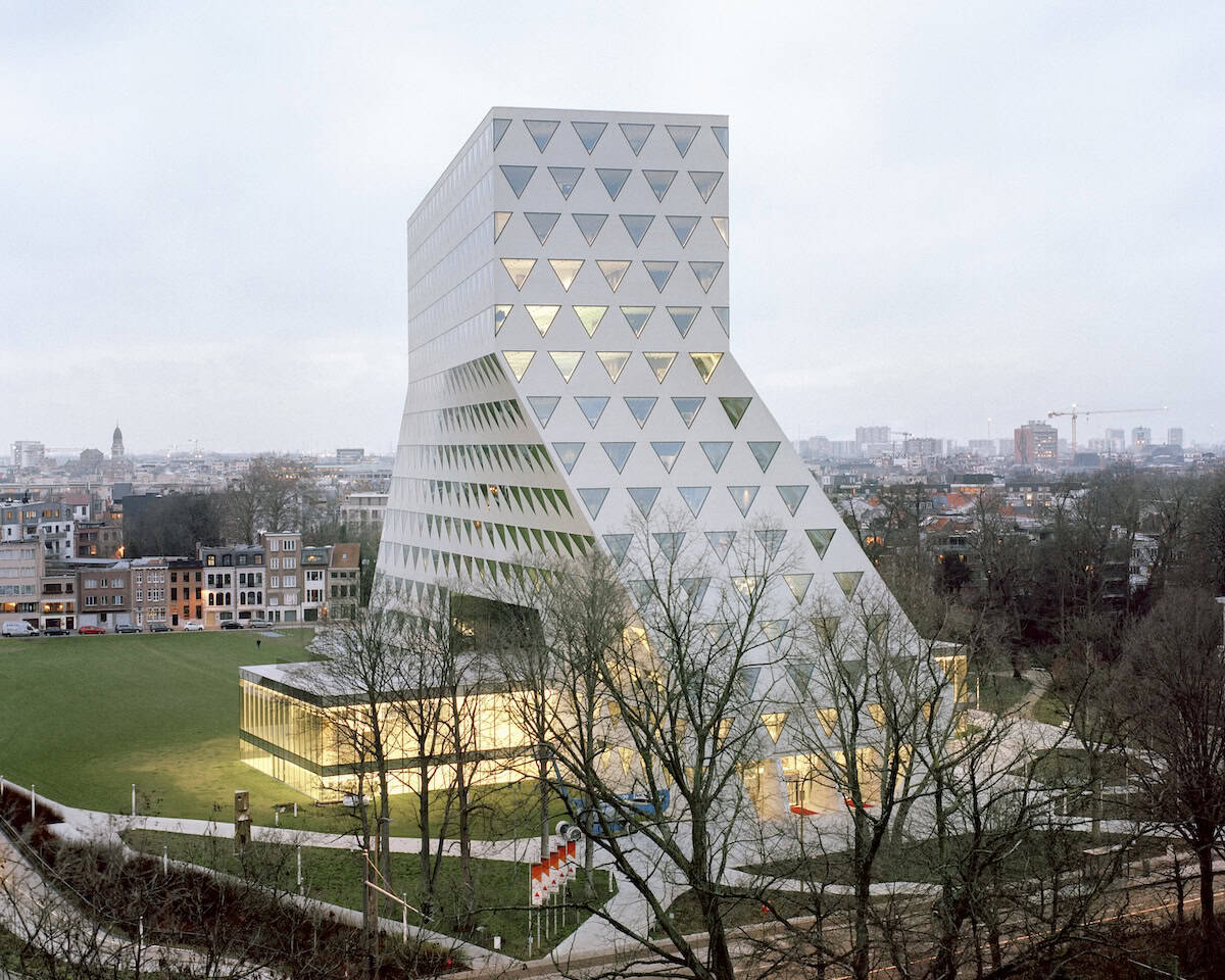 Modern white building with a sloping, pyramid-like base and rows of triangular windows, set in a green park with bare trees and a cityscape in the background.