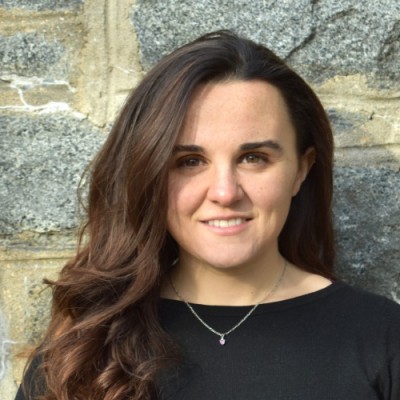 Woman with long wavy brown hair wearing a black top and necklace, standing in front of a stone wall.