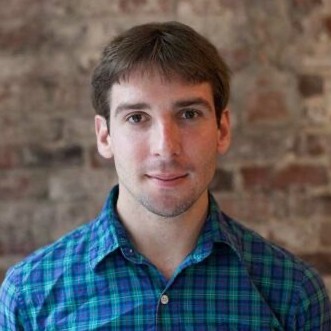 Man with short brown hair wearing a blue plaid shirt, standing in front of an exposed brick wall.