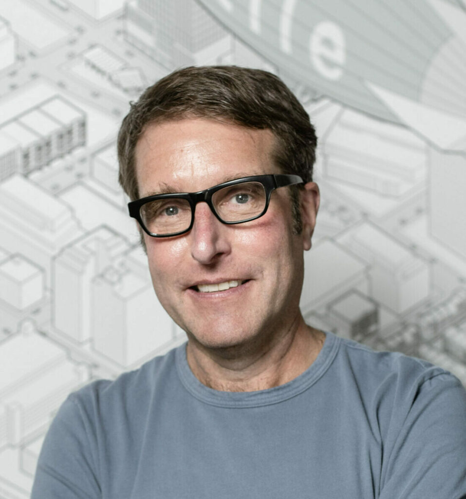 Smiling man with short brown hair and black glasses wearing a gray T-shirt, standing in front of a light architectural-style background.