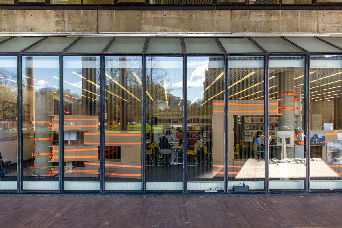 A temporary structure built from cardboard boxes and ratchet straps, seen from outside the windows of Frances Loeb Library.