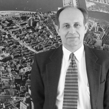 Man in a suit and tie stands in front of an aerial cityscape in a black-and-white photo.