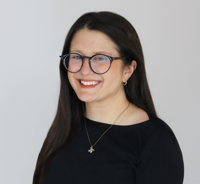 Woman with long dark hair and round glasses smiling at the camera, wearing a black top and a small pendant necklace against a plain light background.
