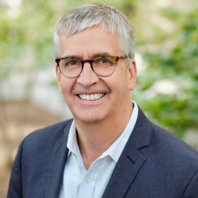 Smiling middle‑aged man with short gray hair and glasses wearing a dark blazer and light shirt, standing outdoors in front of a blurred green background.