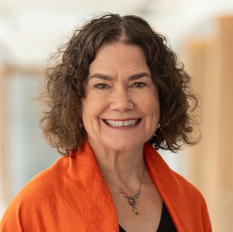 Smiling woman with curly dark hair wearing an orange jacket and colorful necklace in a bright indoor setting.