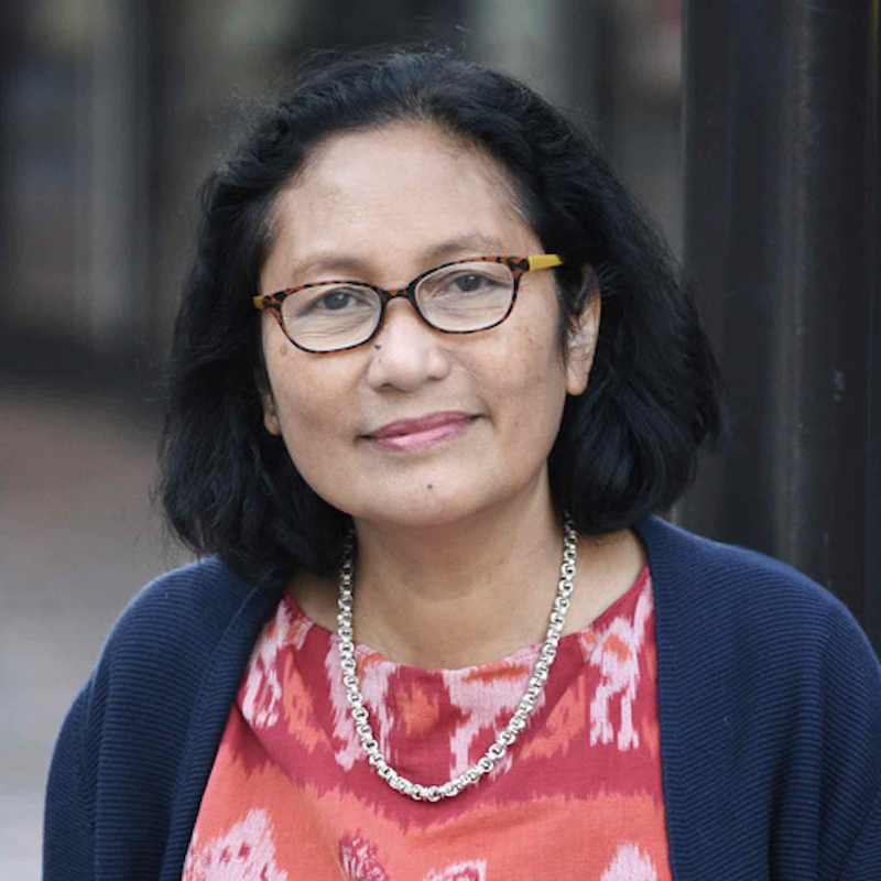 Middle-aged woman with short dark hair and glasses, wearing a patterned red top, blue cardigan, and silver necklace, looking at the camera outdoors.