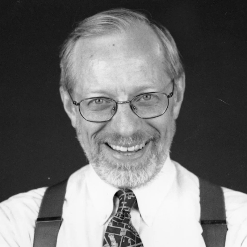 Smiling older man with glasses, beard, and suspenders in a black-and-white portrait.