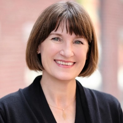 Smiling woman with a short brown bob haircut wearing a black top, standing in front of a softly blurred outdoor background.