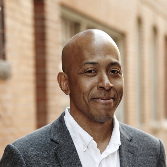 Smiling man with a shaved head wearing a white shirt and gray blazer, standing in a brick alleyway background.