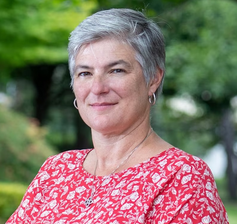 Smiling woman with short gray hair wearing a red patterned top, standing outdoors with greenery in the background.